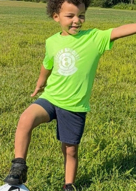 Child in a green shirt playing soccer on a grassy field with trees in the background