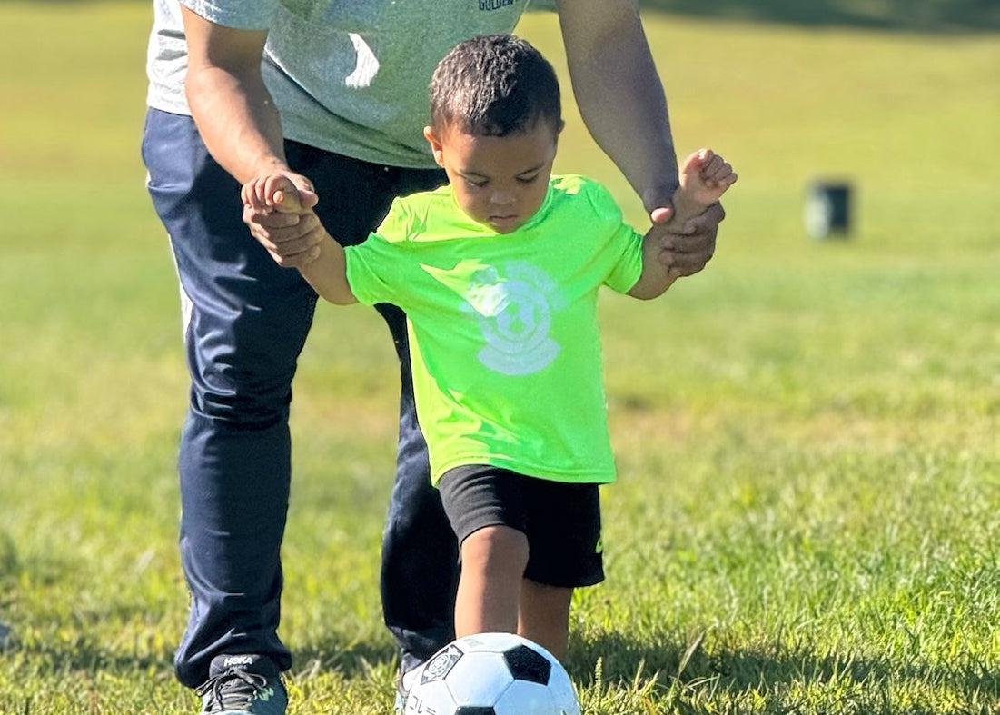 Two children playing soccer on a grassy field with adults nearby.
